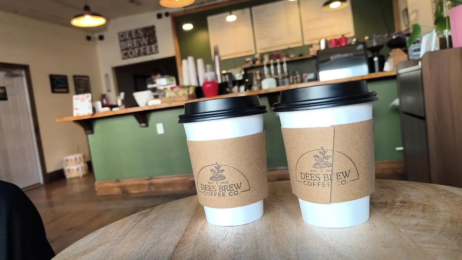 Two Dees Brew cups in kraft sleeves on the table, the counter and menu boards behind them